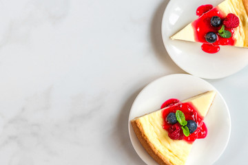 two slices of New York cheesecake in plates with fresh berries, jam and mint on white marble table. top view