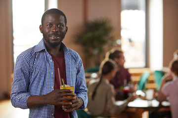 Warm toned waist up portrait of African-American man smiling at camera while holding drink in cafe, copy space