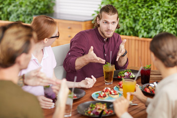 Portrait of handsome young man gesturing actively while talking to friends sitting at table in cafe during holiday reunion, copy space