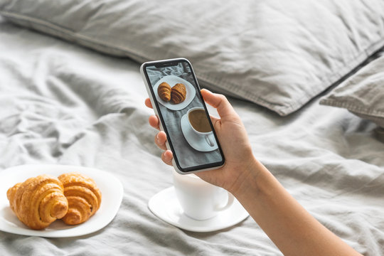 Female hand with mobile phone taking picture of tasty breakfast coffee and croissants in bed, closeup