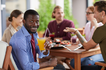 Portrait of African-American man smiling at camera while holding cocktail sitting at table in cafe and enjoying party dinner with friends, copy space