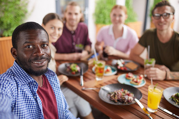 Multi-ethnic group of people posing for selfie photo while enjoying party dinner in cafe together, focus on smiling African-American man in holding camera in foreground, copy space