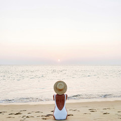 Fototapeta na wymiar Summer holiday fashion concept. Young, tanned woman wearing a beautiful white swimsuit with a straw hat is sitting and relaxing on tropical beach with white sand and watching colorful pink sunset.