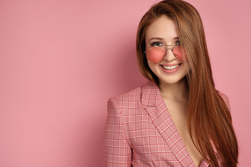 Red-haired girl in a pink jacket stands on a pink background and looks into the camera over his glasses and smiles.