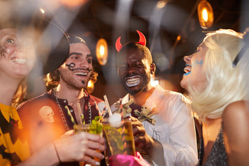 Multi-ethnic group of adult friends wearing Halloween costumes drinking cocktails while enjoying...