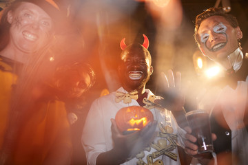 Low angle view at group of adult men wearing Halloween costumes posing during party in club, focus on African-American man holding pumpkin in center