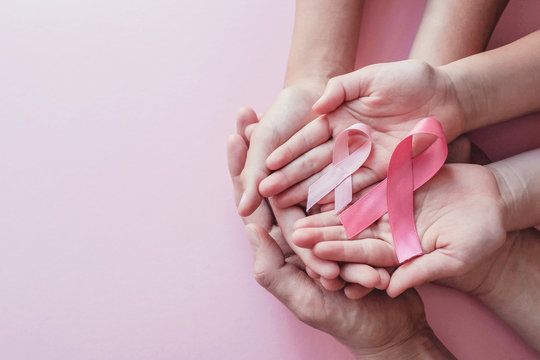 Woman hand holding a light pink ribbon on a pink background - cancer symbol  Stock Photo by wirestock