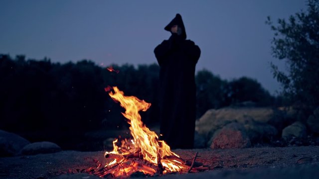 Halloween. Powerful witch or wizard in black costume stands and watches the fire flame in the evening.
