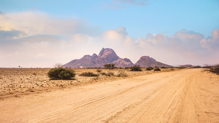 A dirt road running through barren flat plains in the Namib Desert toward the huge granite peaks of Spitzkoppe, Namibia.