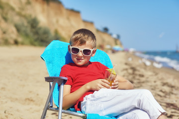 Fototapeta premium a little boy is resting on the sea coast . child sitting on a beach chair holding a glass of juice