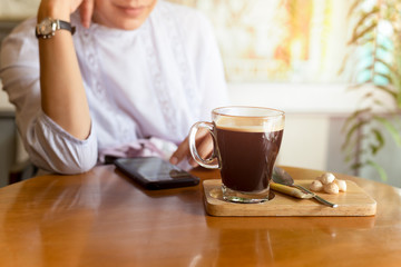 Selected focus coffee cup on table with woman using cell phone in cafe.