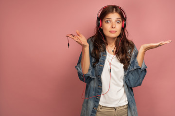 A brunette on a pink background in jeans and headphones is distractedly spreading her arms and holding the wire.