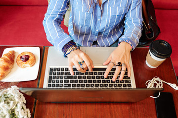 Hands of creative young woman with many rings and bracelets sitting at table with coffee and pastries and working on laptop, view from above