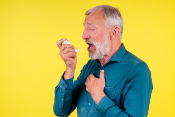Senior man using an asthma inhaler in studio yellow background