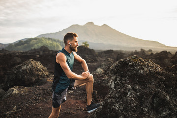 Young hipster runner with beard stretching and warming-up for trail running outdoors. Listening music in air pods. Mountain view on background.