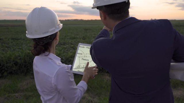 Man and woman discussing future plans for agricultural site with blue prints and hard hat with tablet
