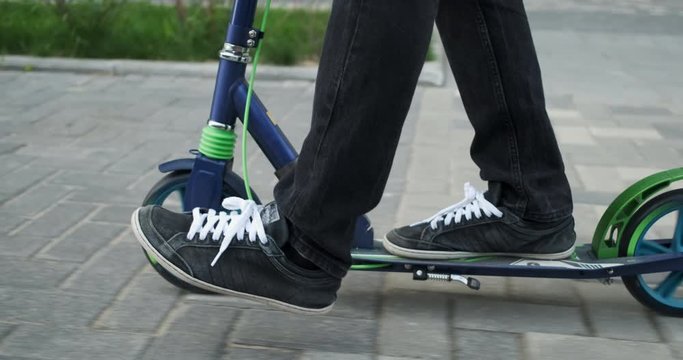 Man in black jeans and black sneakers rides scooter on city sidewalk, summer day