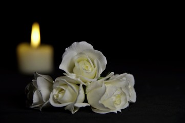 Beautiful White roses with a burning candle on the dark background. Funeral flower and candle on table against black background with copy space. Funeral symbol. Mood and Condolence card concept. 