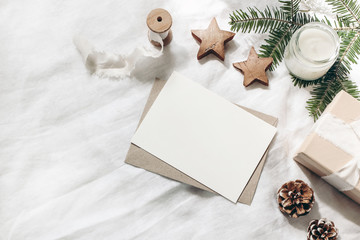 Christmas composition. Blank greeting card, envelope mock-up scene. Decorative corner. Pine cones, fir tree branches, gift box and wooden stars on white table, linen background. Flat lay, top view.