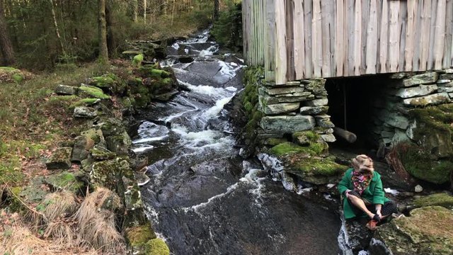 Old woman sitting next to a small creek running in the woods of Sweden during winter time