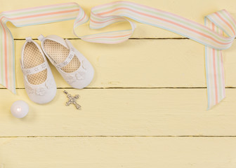 Christening background with white shoes, candle and crystal cross pendant on yellow painted wooden background