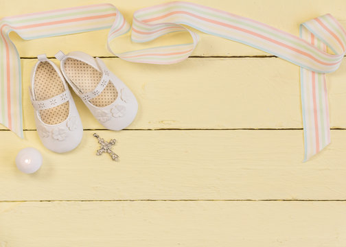 Christening background with white shoes, candle and crystal cross pendant on yellow painted wooden background