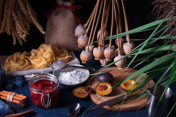 Sweet pasta dessert, noodles with poppy seeds, plum compote from fresh plums, dark background, blue table decorated with fresh flowers. Dry whole poppy plant in background.