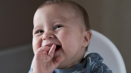 Adorable happy baby boy is looking into the camera. The baby looks around and then smiles and laughs. 1 year old child with brown eyes smiles at home