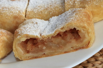 Homemade (home-made) apple strudel with icing sugar from puff pastry (paste) full (filled) pieces of apple with cinnamon on white plate.