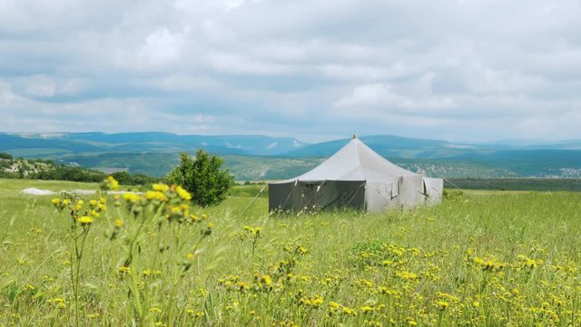 A large canvas military tent stands in the middle of a field in the mountains. In the foreground are yellow wildflowers. In the background mountains and thick gray thunderclouds.