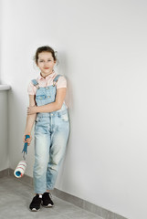 Little girl makes repairs in blue overalls. The child paints the walls. Girl with roller in hands on white wall background.