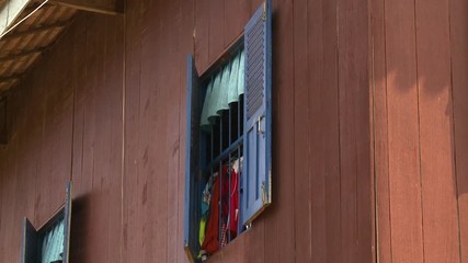 A daylight closeup shot of windows fitted with wooden shutters painted in blue and decorated with a matching light blue short curtain on the second floor of a wooden stilted house with a reddish hue
