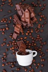 Composition with chocolate, coffee beans and cup. Cup of coffee with grains scattered on black background, top view. Ingredients for delicious drink.