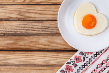 Breakfast on Valentine's Day: fried egg in the shape of a heart on the white plate on the light brown wooden table. Top view.