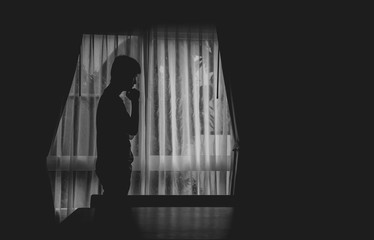 Black and white photo alone man silhouette standing with hand on chin at the window closed with curtains in darkroom. Man stands at window alone, Depression and anxiety disorder concept. .