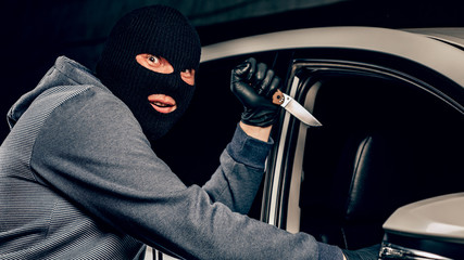 The robber in Balaclava threatening with a knife takes away a bag from the driver of a car. The...