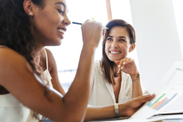 Cheerful happy positive young women colleagues at workplace indoors in office work with paper...