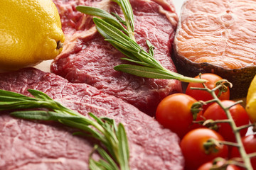 Close up view of raw meat with rosemary twigs near salmon, lemon and branch of tomatoes