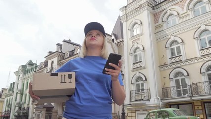 Delivery woman with parcel box using smart phone online map. Low angle shot of a female delivery service courier looking for the house of a client on city street. Shipping concept