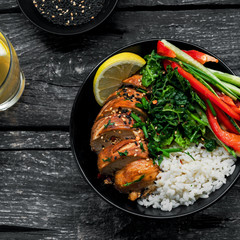 Buddha bowl meal with teriyaki chicken fillet, fresh vegetable salad, stew cabbage and basmati rice on a rustic table. Delicious dish in a black bowl with lemonade. Top view, directly above shot.