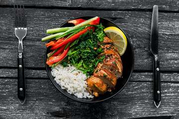Lunch bowl with teriyaki chicken, cucumber, red bell pepper, green onion, stew spinach and basmati rice on a rustic black table. Top view, directly above shot.