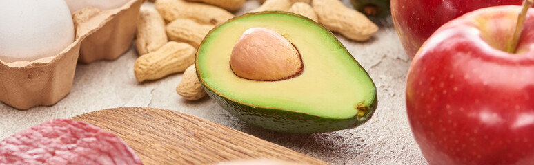 Panoramic shot of half of avocado on marble surface near peanuts, apple and part of egg