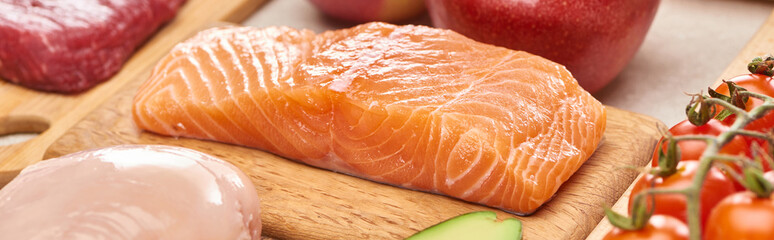 Panoramic shot of raw salmon fillet on wooden cutting board near meat and tomatoes