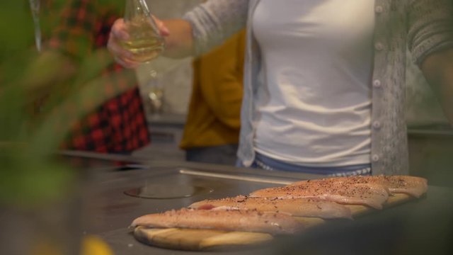 Handheld medium shot of a lady pouring oil on a griddle and spreading at around before placing Saibling Char shish fillets on it with a plant blurred in the foreground