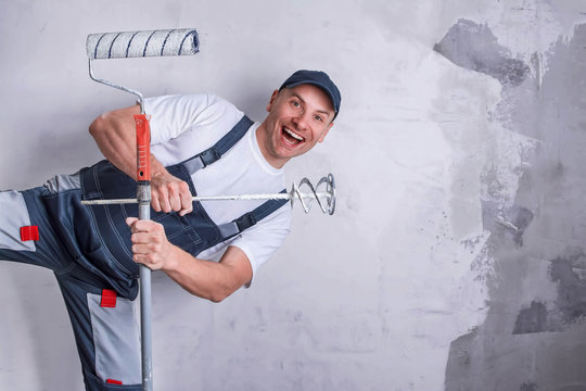 Smiling worker in overall and cap holding a nozzle for electric building mixer covered with putty and a platen for painting in hands. Repair work, construction and profession concept. Copy space