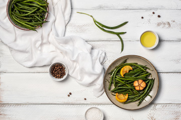 Plate with fried green beans, lemon pieces and garlic, uncooked pods, small bowls with olive oil, sea salt and peppercorns and towel on white wooden table. Healthy food concept. Flat lay, top view