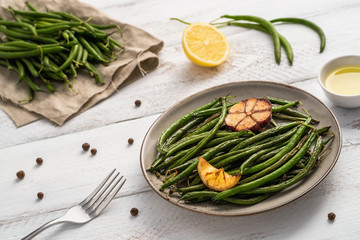 Plate with fried green beans, lemon piece and garlic, uncooked pods on linen napkin, small bowl with olive oil, peppercorns, lemon and a fork on white wooden table. Healthy food concept