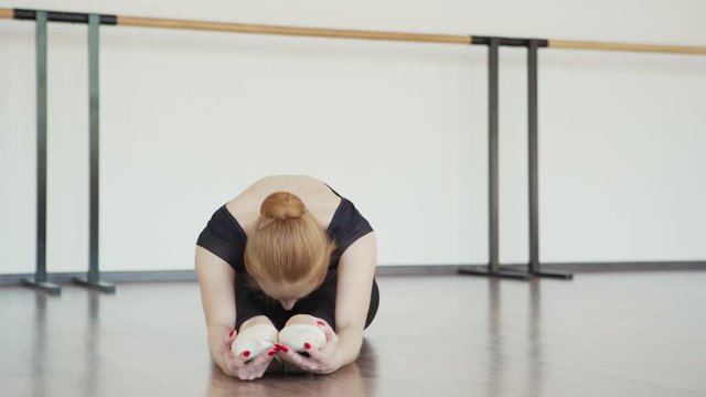 Tracking shot of elegant young ballerina in black leotard and pointe shoes doing forward bend sitting on parquet floor in ballet dance studio