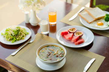 Fresh romantic breakfast table next to window with bread, fruit, juice, milk, noodle soup