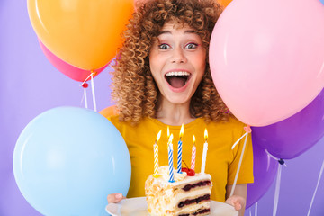 Image of excited young woman celebrating birthday with multicolored air balloons and piece of cake